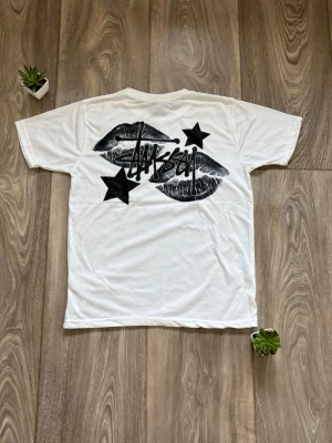 Vit Stüssy t-shirt med tryck - Vit t-shirt från Stüssy i mycket bra skick med svart logga på bröstet och stort tryck med läppar, stjärnor och logga på ryggen. Klassisk passform och korta ärmar. Perfekt för dig som gillar streetwear och vill sticka ut lite extra. Den är ca 64 cm lång och 49 cm bred