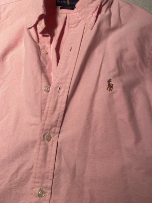 Rosa skjorta från Ralph Lauren - Säljer en snygg rosa skjorta från Ralph Lauren i slim fit-modell. Skjortan har klassisk krage, knappar framtill och den ikoniska broderade loggan på bröstet. Perfekt för dig som gillar stilrena och tidlösa plagg.