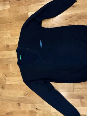 Mörkblå V-ringad tröja Benetton stl L - Klassisk mörkblå V-ringad tröja från Benetton. Tröjan har långa ärmar och en liten bröstficka med kontrastfärgad detalj. Perfekt för lager på lager och enkel att matcha med olika stilar.