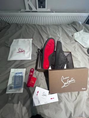 Christian Louboutin svarta sneakers - Exklusiva svarta sneakers från Christian Louboutin med ikonisk röd sula och diskret logga på sidan. Skorna har en klassisk låg siluett, snörning framtill och är tillverkade i mocka. Kommer med originalkartong, dustbag och certifikat.