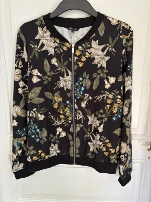 Svart tröja med dragkedja och blommigt mönster - Snygg svart tröja/ mycket tunn jacka med dragkedja och  blommönster i vitt, grönt, blått och gult.  Ribbade muddar vid hals, ärmslut och nederkant. 