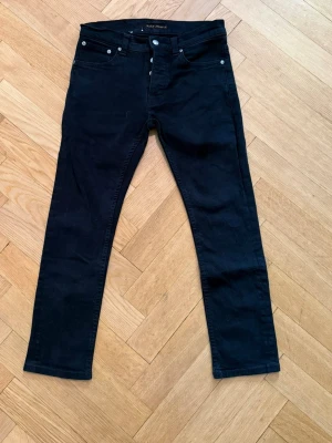 Svarta jeans från Nudie - Svarta jeans från Nudie med klassisk femficksdesign och knappgylf. Modell: Grim Tim. Storlek: 30/30. Jeansen har raka ben och är tillverkade i ett stretchigt denimtyg för extra komfort. Perfekta för en clean och stilren look.