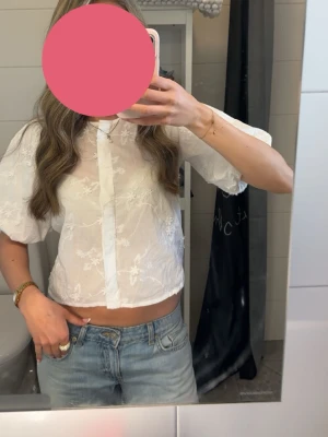 Vit blommig blus med puffärm - Söt vit blus med broderade blommor och puffiga korta ärmar. Blusen är lätt transparent och har en rak passform med dold knäppning framtill. Perfekt för en trendig och fräsch look.