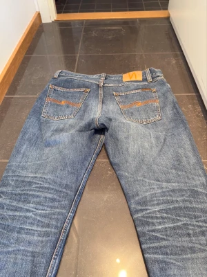 Blå jeans från Nudie Jeans - Säljer ett par klassiska blå jeans från Nudie Jeans med raka ben och snygga slitningar. Jeansen har två bakfickor med Nudie-logga i orange och är gjorda i slitstarkt denim. Perfekta för en avslappnad och trendig look. Aldrig använda! Nypris: 1600 kr