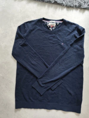 Mörkblå stickad tröja Tommy Hilfiger - Mörkblå stickad tröja från Tommy Hilfiger med rund halsringning och diskret logga broderad på bröstet. Tröjan har långa ärmar och ribbade muddar vid ärmslut och nederkant. Perfekt för en stilren och klassisk look.