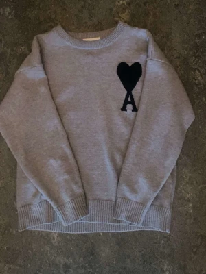Ami Paris - Säljer en grå stickad tröja med svart hjärta och bokstaven A på bröstet. Tröjan har rund halsringning, ribbade muddar och nederkant. Perfekt för dig som gillar enkel men snygg design.