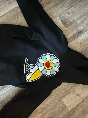 Svart OVO x Murakami hoodie med blomma - Säljer en svart hoodie från October's Very Own x Murakami med stor broderad blomma i blått, vitt och gult framtill. Hoodien har huva och är tillverkad i mjukt material. Perfekt för dig som gillar streetwear och unika samarbeten.