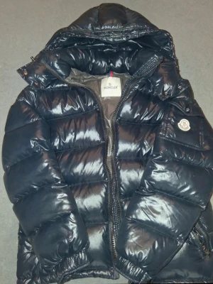 Mörkblå dunjacka från Moncler - Snygg svart dunjacka från Moncler med glansig finish och klassisk puffer-design. Jackan har huva, dragkedja framtill och Moncler-logga på ärmen. Perfekt för kalla dagar när du vill hålla stilen och värmen. Storlek 3/L men passar mer som M.