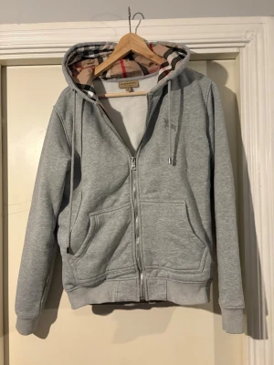 Grå zip-hoodie från Burberry - Snygg grå hoodie med dragkedja från Burberry. Klassisk huva med det ikoniska rutmönstret på insidan, dragsko och två stora fickor framtill. Mjuk bomullsblandning och ribbade muddar vid ärmslut och nederkant. Perfekt för en chill och stilren look.