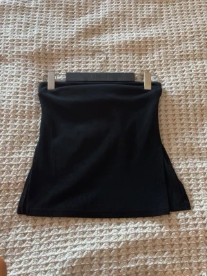 Svart ribbad tubtopp med slits - Svart ribbad tubtopp i stretchigt material med en liten slits på sidan. Enkel och stilren design som passar perfekt till jeans eller kjol. Sitter tight och är axelbandslös för en clean look.