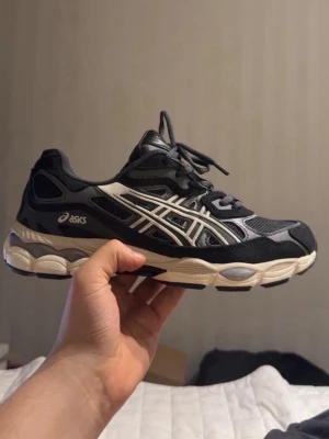 Svarta ASICS sneakers med vita detaljer - Snygga svarta ASICS sneakers med vita och grå detaljer. Skon har en sportig design med mesh- och syntetmaterial, samt en vit och svart sula som ger en cool kontrast. Perfekt för dig som gillar streetwear och sportig stil.