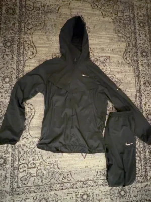 Nike Tracksuit Uv Repel - Säljer ett svart träningsset från Nike med jacka och byxor. Jackan har huva, dragkedja och reflekterande detaljer samt Nike-logga på bröstet. Byxorna har resår i midjan och liten Nike-logga. Perfekt för löpning eller träning utomhus. Köpt för 1600 använd 10 gånger max