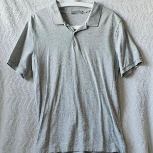 Grå pikétröja från Calvin Klein Jeans - Snygg grå pikétröja från Calvin Klein Jeans med klassisk krage och korta ärmar. Tröjan har knappar vid halsen och är tillverkad i mjukt bomullsmaterial. Perfekt för en stilren och avslappnad look.