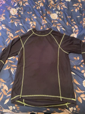 Svart långärmad träningströja Bluewear - Svart långärmad träningströja från Bluewear med synliga gröna sömmar och diskret ribbad struktur. Perfekt för träning eller utomhusaktiviteter. Tröjan har rund hals och är tillverkad i ett syntetmaterial som andas.