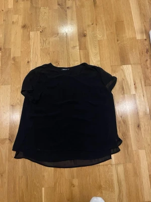 Svart kortärmad blus Gina Tricot stl 42 - En enkel och stilren svart blus från Gina Tricot. Blusen har rund halsringning, korta ärmar och är något längre baktill. Tillverkad i ett lätt och tunt material som ger ett snyggt fall.