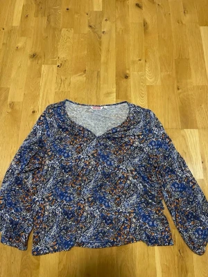 Mönstrad blus från Indiska stl L - Blus från Indiska med blått, orange och vitt paisleymönster. Långärmad modell med rundad halsringning och lätt rynkning framtill. Tunt och mjukt material som känns skönt mot huden. Perfekt för dig som gillar färg och mönster.