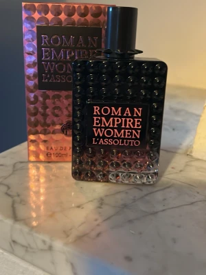 Roman Empire Women L'Assoluto parfym - Roman Empire Women L'Assoluto är en Eau de Parfum i en svart och rosa skimrande flaska med nitar och elegant design. Flaskan rymmer 100 ml och har en lyxig känsla med matchande förpackning. Perfekt för dig som gillar exklusiva dofter och snygga flaskor.