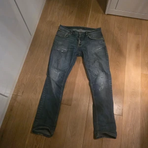 Nudie jeans - Säljer ett par blå jeans med slitningar och hål på flera ställen för en cool, avslappnad look. Klassisk femficksmodell med rak passform. Perfekta för dig som gillar en trendig och edgy stil. Materialet är denim i bomull. Det syns att de är ancända och reparerade några gånger,det finns två jål men som man enkelt kan lämmna in gör reperation vid sin lokala nudie jeans reparationsbutik