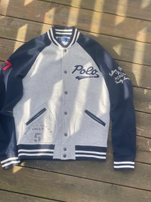 Blå och grå bomberjacka Polo Ralph Lauren - Snygg bomberjacka från Polo Ralph Lauren i blått och grått med broderade detaljer, tryck och randiga muddar. Jackan har knappar framtill, två fickor och broderad text på ärmen samt siffran 67 på axeln.