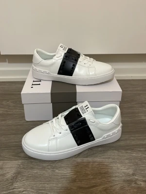 Valentino sneakers  - Snygga vita sneakers från Valentino Garavani med en bred svart rem över vristen dekorerad med nitar. Skorna har en klassisk låg profil, rund tå och är tillverkade i slätt läder. Perfekta för dig som vill sticka ut med stilrena detaljer.