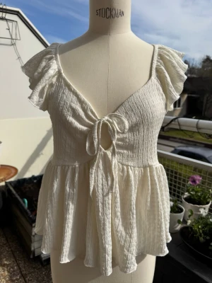 Vit blus med volangärmar och knyt - Supersöt vit blus med volangärmar och v-ringning. Toppen har knyt framtill och är tillverkad i ett lätt, krinklat bomullstyg som ger en luftig känsla. Perfekt för varma dagar och ger en romantisk vibe till din outfit. Lager 157