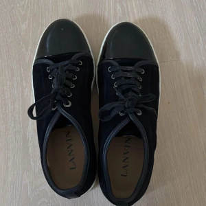 Svarta sneakers från Lanvin med lackad tå - Snygga svarta sneakers från Lanvin med klassisk snörning och vit sula. Skorna har en unik design med lackad tå i svart och ovandel i mocka. Perfekta för dig som vill ha en stilren men ändå cool look.