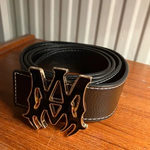 Amiri bälte - Säljer ett svart Amiri bälte i skinn med grov textur och vita sömmar längs kanterna. Spännet är stort, guldig-design med svarta detaljer. Perfekt för att ge outfiten en unik och edgy touch. Helt nytt! 