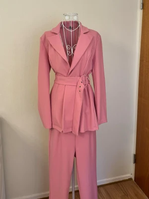 Ny rosa kostym set - Snygg rosa kostym med kavaj och matchande byxor. Kavajen har ett brett bälte i midjan och klassisk krage. Oanvänt och i ny skick. Storlek S. 