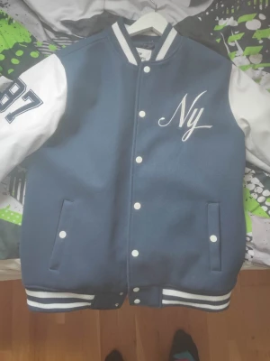 Blå bomberjacka New Garments - Snygg blå bomberjacka från New Garments med vita ärmar, broderad text 'Ny' på bröstet och siffran 87 på ena ärmen. Jackan har vita knappar, randiga muddar och två fickor framtill. Perfekt för en sportig och trendig look.