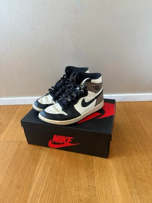 Nike Air Jordan 1 High Mocha  - Säljer ett par Nike Air Jordan 1 High i svart, vitt och brunt läder med klassisk svart swoosh och högt skaft. Ikonisk vinge-logga på sidan och svart yttersula. Perfekta sneakers för dig som gillar streetwear och vill sticka ut med en tidlös modell.