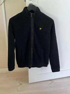  Lyle & Scott - Mysig svart teddycardigan från Lyle & Scott med dragkedja framtill och gul logga på bröstet. Hög krage och två fickor. Perfekt för lager på lager och en chill look.
