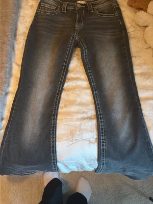Blå bootcut jeans med slitningar - Säljer ett par blå bootcut jeans med snygga slitningar och kontrastsömmar. Jeansen har klassiska fem fickor och detaljerade bakfickor med V-formad söm. Perfekta för dig som gillar retrostil och vill ha ett par jeans med lite vidare ben.