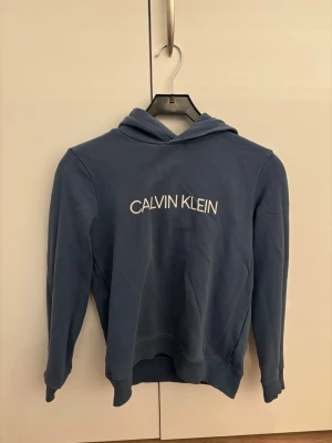 Blå hoodie från Calvin Klein - Snygg blå hoodie från Calvin Klein med vit logga tryckt på bröstet. Hoodien har huva, långa ärmar och ribbade muddar vid ärmslut och nederkant. Perfekt för en avslappnad och trendig stil.
