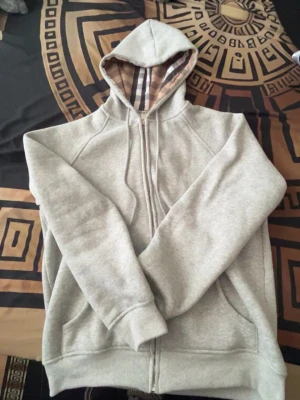 Burberry Hoodie - Snygg grå zip-hoodie med klassisk dragkedja och fickor framtill. Insidan av huvan har ett rutigt mönster i beige, svart, vitt och rött som ger en extra touch. Perfekt för en chill och avslappnad stil. Mjuk bomullskänsla och ribbade muddar.