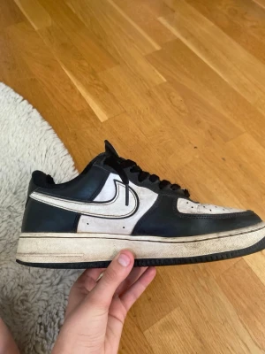 Nike Air Force 1 svart/vit sneaker - Klassisk Nike Air Force 1 sneaker i svart och vitt läder med svart sula och vita detaljer. Lågt skaft och tydlig Nike-logga på sidan samt AIR-brodyr på hälen. Perfekt för dig som gillar streetwear och tidlös design.