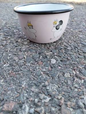 Mumin-skål i ljusrosa emalj - Söt ljusrosa emaljskål med svart kant och motiv av Muminfigurer. Perfekt för snacks eller frukost, med klassisk Mumin-design och rund form. Skålen har vit insida och är lätt att rengöra.