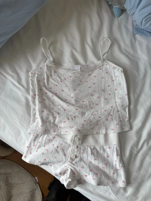 Vitt ribbat pyjamasset med små rosa blommor - Sött pyjamasset i vitt med ribbad struktur och små rosa blommor. Setet består av ett linne med tunna axelband och matchande shorts med knappar framtill. Perfekt för mysiga hemmakvällar. Mjukt och bekvämt material.