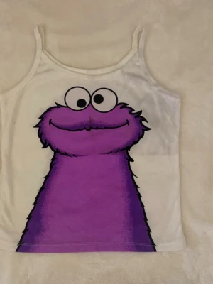 Vit linnetopp med lila monstertryck - Säljer en vit linnetopp med smala axelband och ett stort tryck av ett lila tecknat monster på både fram- och baksidan. Toppen har en lekfull och färgglad design, perfekt för dig som gillar unika plagg med humor. Fläckarna som finns (kolla bilder) kommer säkert/troligtvis försvinna i tvätten men det har blivit avfärgat från jeans i lådan de vart i. 