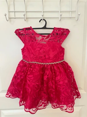 Rosa klänning med pärlbälte - Säljer denna supersöta rosa bebis klänning med blommig spets och transparenta detaljer upptill. Klänningen är köpt från Rare Editions i USA. Klänningen har ett glittrande pärlbälte i midjan och volangig kjol. Passar storlek 1-2 år🫶🏼