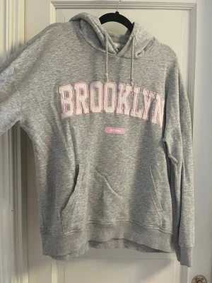 Grå hoodie BROOKLYN tryck  - Grå hoodie med Rosa tryck BROOKLYN. Använt några gånger. Fläck på magen! Köpt på Stadium