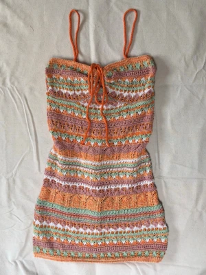 Färgglad virkad kortklänning - Säljer en unik virkad kortklänning med smala axelband och snörning framtill. Klänningen har ett färgglatt randigt mönster i orange, grön, vit och rosa. Använd 1 gång. 🏖️