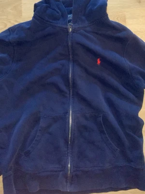Mörkblå hoodie från Polo Ralph Lauren - Snygg mörkblå hoodie från Polo Ralph Lauren med dragkedja och klassisk röd logga på bröstet. Hoodien har huva, känguruficka och är tillverkad i mjukt bomullsmaterial. Perfekt för en avslappnad och stilren look. Står 14-16 år skulle säga att den sitter bra på 155-166