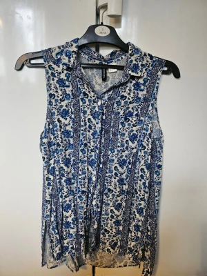 Blommig ärmlös blus i blått och vitt H&M - Säljer en ärmlös blus med blått och vitt blommönster. Blusen har krage, knappar framtill och en lös passform. Perfekt för varma dagar och enkel att styla med jeans eller kjol.