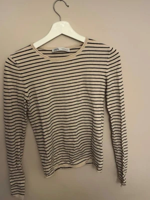 Randig långärmad topp från Zara - Säljer en beige långärmad topp från Zara med smala svarta ränder. Toppen har rund halsringning och är i ett mjukt, stretchigt material som sitter snyggt på kroppen. Perfekt att styla med jeans eller kjol.