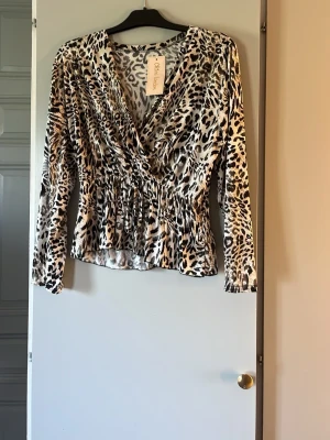 Leopardmönstrad omlottblus från Sisters Point - Trendig leopardmönstrad omlottblus med smockad midja och långa ärmar. Blusen har djup v-ringning och är tillverkad i ett glansigt, mjukt material som ger en lyxig känsla. Perfekt för ragge! Storlek M/L men är lite mindre i storleken 
