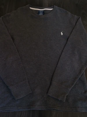 Mörkgrå tröja från Polo Ralph Lauren - Säljer en mörkgrå stickad tröja från Polo Ralph Lauren med klassisk rund hals och långärmad passform. Tröjan har en vit broderad logga på bröstet och är tillverkad i mjukt stickat material. Axelbredd: 48cm / Längd: 62cm. Nypris: 995kr / Mitt pris: 349kr