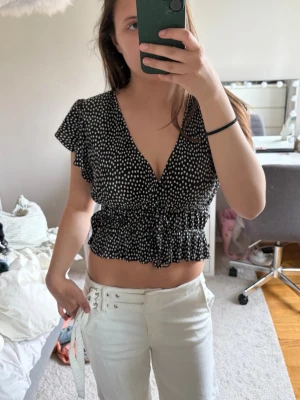 Svart prickig croppad blus - Svart croppad blus med vita prickar och djup v-ringning. Toppen har volangärmar och resår i midjan för en snygg passform!