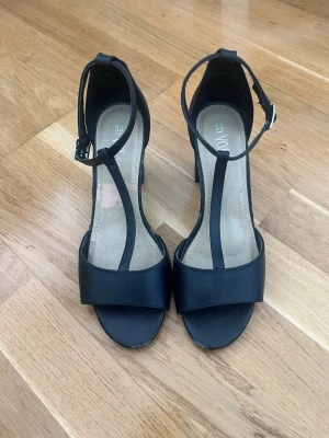 Svarta T-rem pumps från VOX - Snygga svarta pumps från VOX med T-rem och öppen tå. Skorna har en justerbar ankelrem med spänne och en stadig klack. Tillverkade i ett slätt material som ger en stilren look. Perfekta för dig som gillar klassisk och trendig stil.