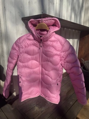 Rosa dunjacka med huva från Peak Performance NY ALDRIG ANVÄND  - Snygg rosa dunjacka från Peak Performance med vågigt quiltat mönster och praktisk huva. Jackan har dragkedja framtill och två fickor med dragkedja. Perfekt för kalla vinterdagar och riktigt skön att bära.