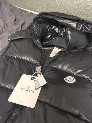 Svart Moncler väst - Snygg svart Moncler väst med glansig finish, dragkedja framtill och två fickor med dragkedja. Västen har avtagbar huva och klassisk Moncler-logga på bröstet. Perfekt för lager-på-lager och streetstyle. Västen har NFC-tag och är 1:1. Passar L storlek